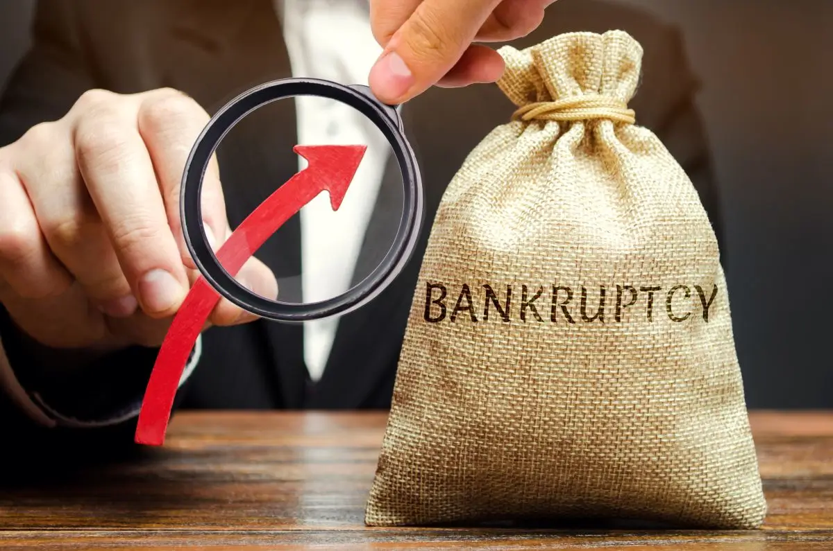 Businessperson holding a magnifying glass with an upward red arrow next to a burlap bag labeled ‘Bankruptcy,’ symbolizing financial evaluation and bankruptcy qualification.