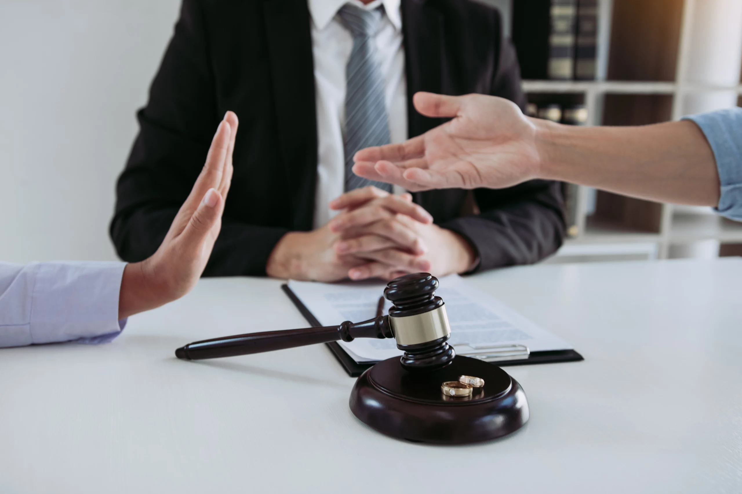 Two hands gesture in disagreement over a gavel and wedding rings, with a suited person in the background, symbolizing a contested divorce in Missouri.