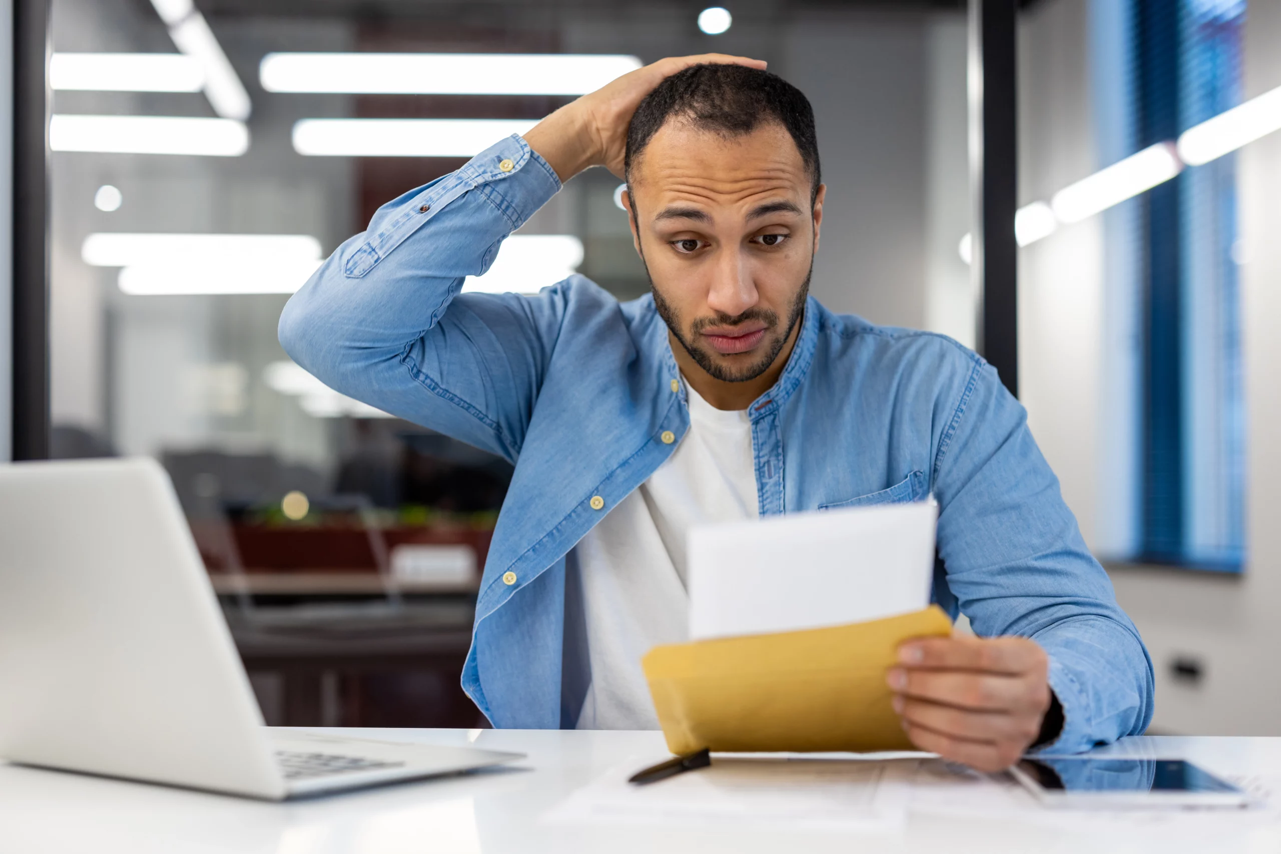 Worried man in a blue shirt reads a letter at his desk with a laptop, showing stress over a possible debt collection lawsuit before going to court.