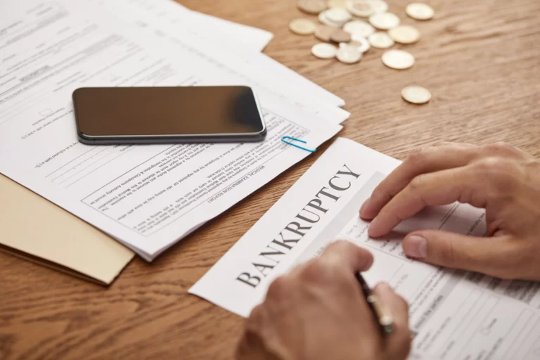 Person signing bankruptcy paperwork with coins, documents, and a phone on the table, representing the costs and financial considerations of filing for bankruptcy in Missouri