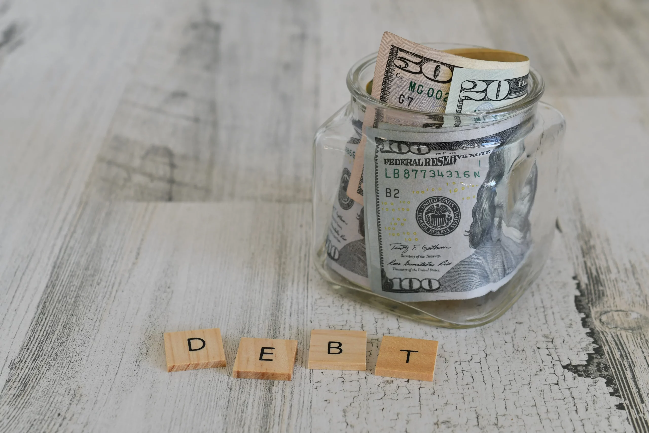 Glass jar filled with assorted U.S. dollar bills on a rustic wooden surface, arranged beside wooden letter tiles spelling “DEBT,” symbolizing financial burden and non-dischargeable obligations under Chapter 7 bankruptcy law.